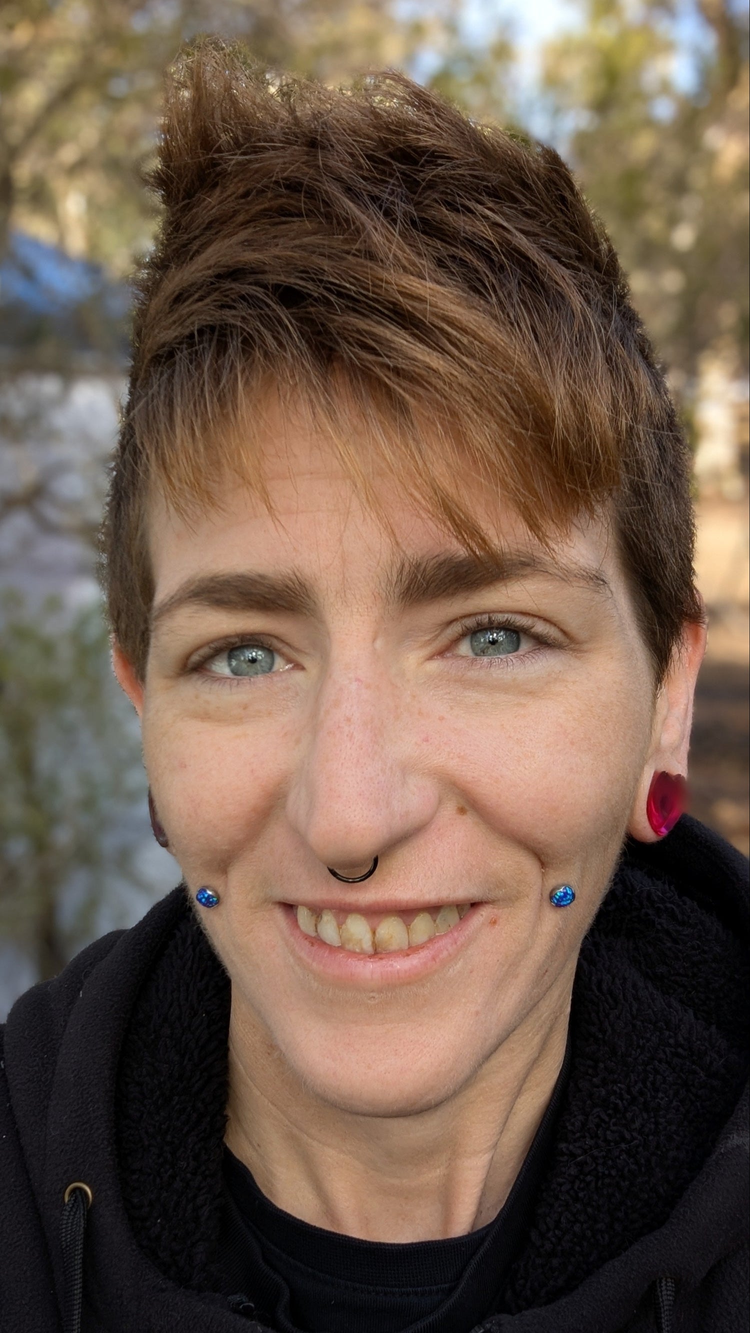 Person with short hair and piercings standing outdoors with trees in the background